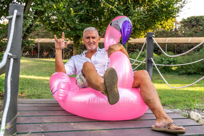 Smiling handsome middle aged man making sign of the horns sitting on an inflatable pink flamingo toy