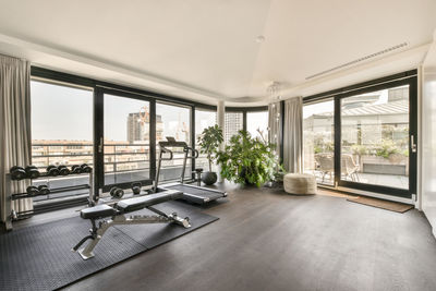 Interior of spacious hall with treadmill and sports supplies in front of large panoramic window overlooking cityscape in modern luxurious apartment