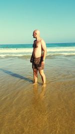 Full length of shirtless mid adult man standing at beach against clear sky