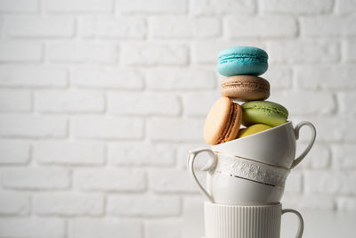 Stack of white coffee cups filled with macaroons on the edge of the table, white brick wall backgro