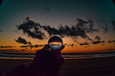 Man holding sunglasses at beach against sky during sunset