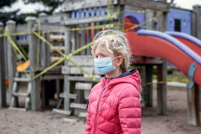  girl in face mask standing on closed playground. kids play area locked with yellow caution tape