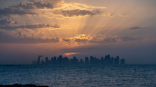 Silhouette buildings by sea against sky during sunset