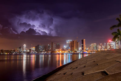 Illuminated buildings by river against sky at night