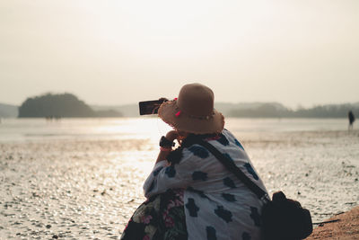 Rear view of woman crouching and photographing from smart phone against sky