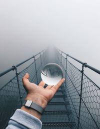 Cropped hand of man holding crystal ball on footbridge against fog