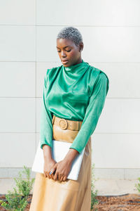 Serious young african american businesswoman with short dyed hair in fashionable outfit standing on street with laptop in hand and looking down