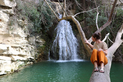 Woman looking at waterfall while sitting on tree trunk in forest