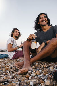 Smiling friends with alcohol bottles sitting on pebbles at beach against sky