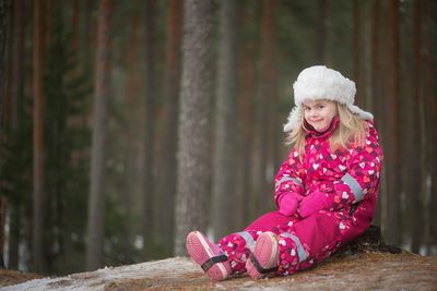 Portrait of smiling girl in warm clothing sitting by trees in forest during winter