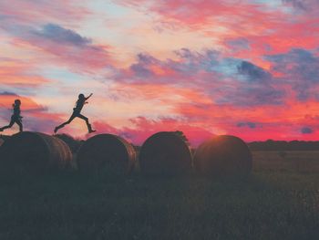 Silhouette friends running on hay bales against cloudy sky at sunset