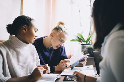 Teenage students discussing over book while sitting together at table in house