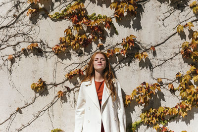 Young redhead woman wearing jacket standing with eyes closed against ivy wall