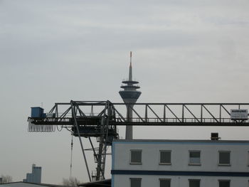 Low angle view of crane by building with rheinturm tower in background during sunset