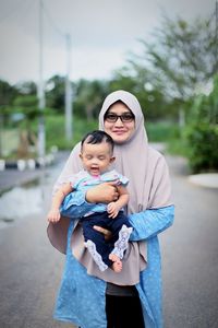 Portrait of smiling mother carrying cute daughter while standing on road against sky