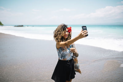 Woman taking selfie while standing at beach against sky