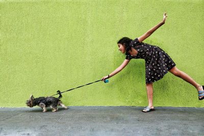 Full length of girl walking behind dog against green wall