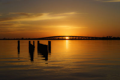 Silhouette wooden posts in sea against orange sky