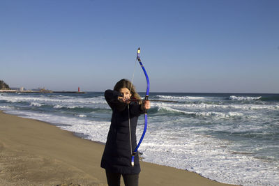 Female archer aiming while standing against sea at beach