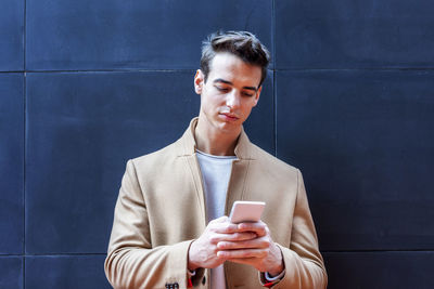 Young man using phone while standing against wall