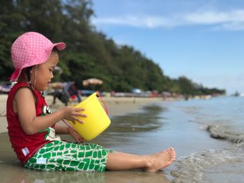 Side view of girl holding bucket while sitting at beach