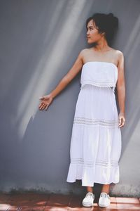 Beautiful young woman wearing white dress while standing against wall