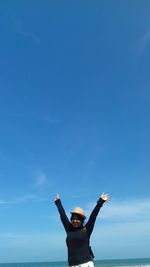 Teenage girl with arms raised standing by sea against blue sky during sunny day