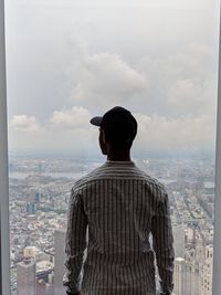 Rear view of man looking at cityscape through window against sky