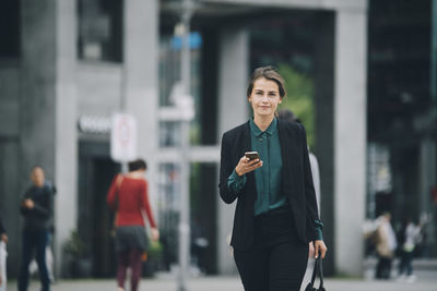 Portrait of confident businesswoman holding smart phone while walking on street in city