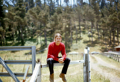 Portrait of young man sitting on fence against trees