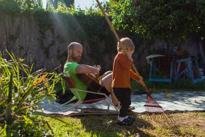 Man playing guitar while looking at son cleaning yard