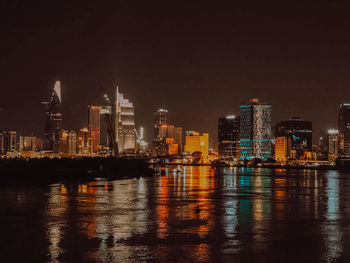 Illuminated buildings by river against sky at night