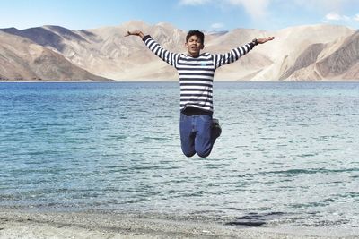 Boy jumping on lakeshore against mountains