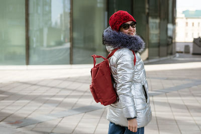 Happy young woman in winter jacket with backpack near building
