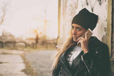 Fashionable young woman wearing leather jacket using smart phone against wall