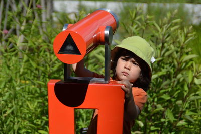 Boy using red hand-held telescope against plants on sunny day