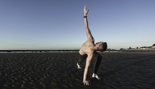 Full length of shirtless man exercising at beach against clear sky