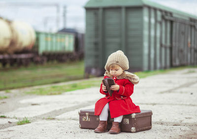 Charming baby in a red coat with a suitcase is waiting for a train on the platform