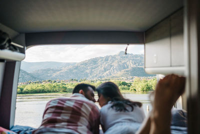 Back view of positive hispanic couple of travelers wrapped in plaids drinking beverages and chilling near pond in summer while looking at each other inside caravan