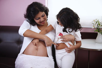 Girl and pregnant mother touching each other belly at home