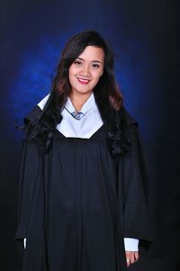 Portrait of smiling young woman wearing graduation gown standing against blue background