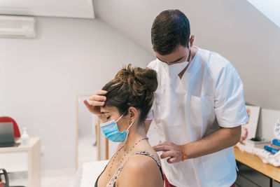 High angle of male physiotherapist massaging back of calm female patient in mask sitting on medical table in modern clinic