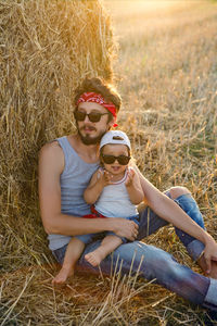 Father and son in t-shirts sitting next to a haystack on a sloping field during sunset