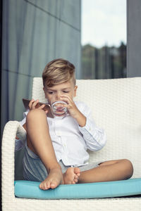 Full length of boy drinking water while using smart phone on sofa at balcony