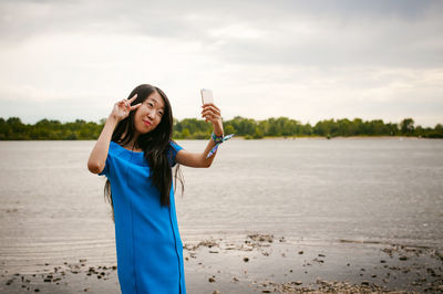 Woman taking selfie with smart phone by lake against sky