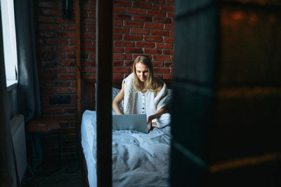 Adult woman freelancer with blonde long hair in casual clothes using laptop on bed at home