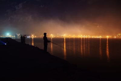 Silhouette cityscape by sea against sky at night