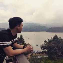 Young man looking at lake against cloudy sky