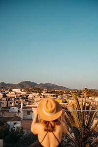 Back view of unrecognizable young female tourist in stylish black swimsuit and straw hat standing on rooftop and admiring town against cloudless blue sky in mallorca