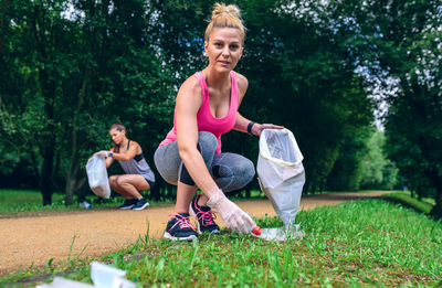 Full length of happy women picking up garbage while crouching on grass against trees